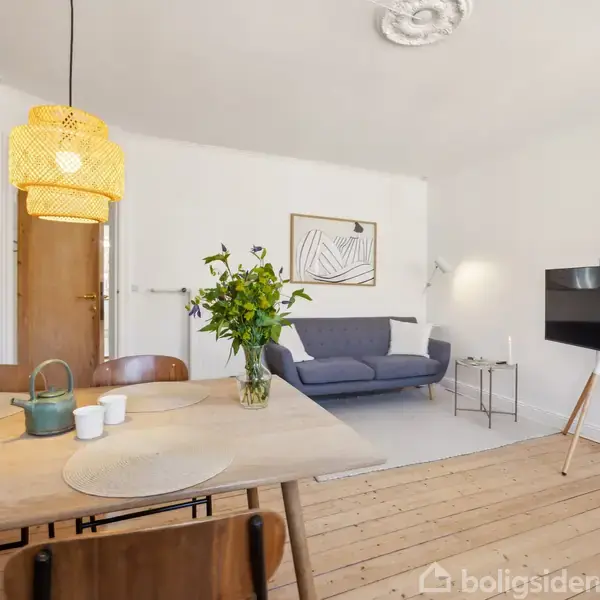 En stue med en blå sofa står ved en hvid væg med kunst. En lampe hænger over et spisebord med en grøn tekande og blomster. En fladskærm er på et stativ.