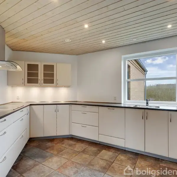 Et moderne køkken med hvide skabe og mørk bordplade, set i dagslys gennem et stort vindue. Skiferlignende gulvfliser og indbyggede loftslamper tilfører en lys, rummelig atmosfære.