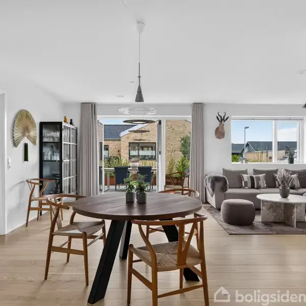 Rundt spisebord med stole, stillestående i moderne stue, omgivet af sofa, glasvitrine og store vinduer, der lader naturligt lys ind. En abstrakt vægdekoration hænger nær en åben dør til et soveværelse.