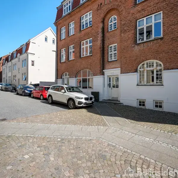 En række biler, herunder en sølvfarvet bil, holder parkeret langs en brostensbelagt gade foran en murstensbygning med store vinduer.