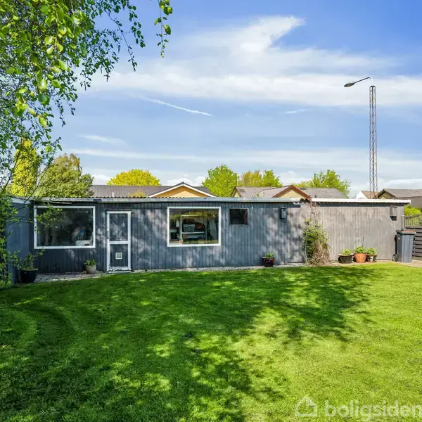 En grå bygning med vinduer står i solen. Foran er der en velholdt græsplæne omkranset af et hegn med træer og buske. Skyfri himmel i baggrunden.