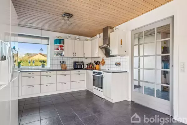 Et lyst, moderne køkken med hvide skabe og grå flisegulve. En ovn og emhætte er centralt placeret, omgivet af køkkenredskaber. Et vindue viser udsigt til en have.