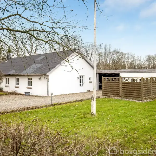 Hvidt hus med grå tag, omgivet af græsplæne og træer, står ved siden af grusindkørsel og skur med træhegn, i landlig omgivelser.
