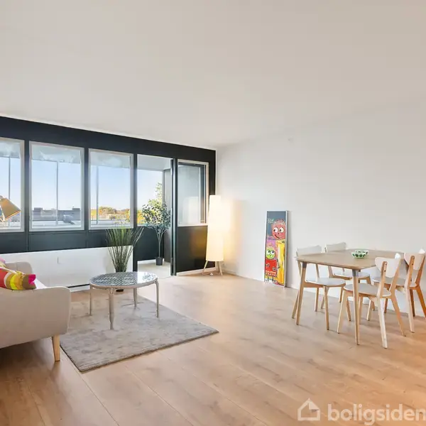 Sofaen står i en stue med lyse trægulve, ved siden af et sofabord og en gulvlampe. Store vinduer giver udsigt til det udendørs. En spiseplads er i hjørnet.