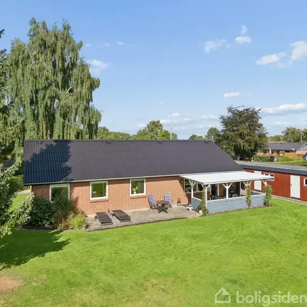 Hus med sort tag står i solskin. Have med græs og store træer omgiver huset. Terrassen har havemøbler, og en carport er til højre med en bil parkeret.