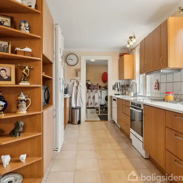 Et køkken med træskabe og åbne hylder viser familiefotos og keramik. Komfuret er placeret langs højre væg. En døren i enden fører til et bryggers.