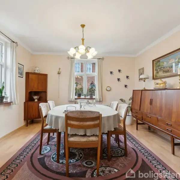 Et rundt bord med dug står i midten af en spisestue med lyse vægge og træmøbler. Et lysekrone hænger over bordet, omkranset af stole. Vinduet har gardiner.