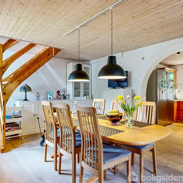 Træ spisebord med seks stole, dekoreret med en vase blomster, står i spisestue. Trætrappe fører op til næste etage; sort pendellampe hænger over bordet. Køkken ses gennem buegang.