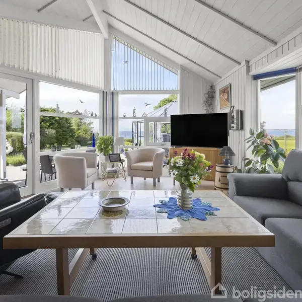 Lyst stueområde med sofagruppe og sofabord, udsigt til terrasse. Store vinduer giver udsigt til haven og havet. Rummets vægge og loft er hvide med synlige bjælker.