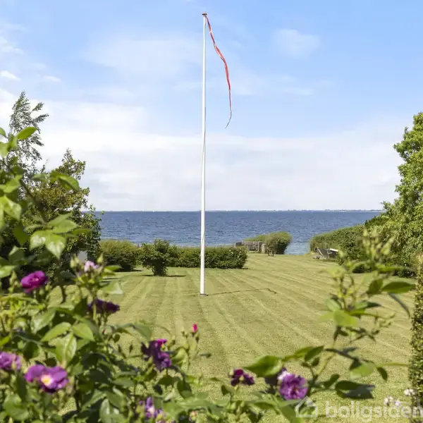 Flagstang med vimpel står stille i en velholdt græsplæne, omkranset af buske og blomster, med udsigt over en rolig havstrækning under blå himmel.