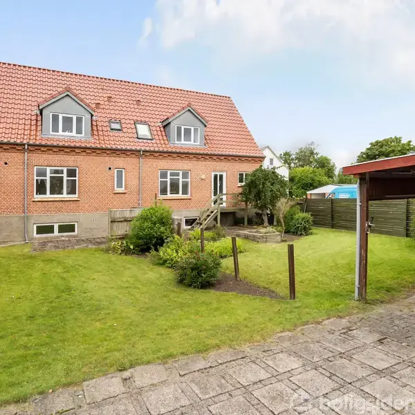 Rødt murstenshus står i en grøn have med græsplæne. Til højre holder en orange bil under en carport. Baggrunden viser en klar himmel og få træer.