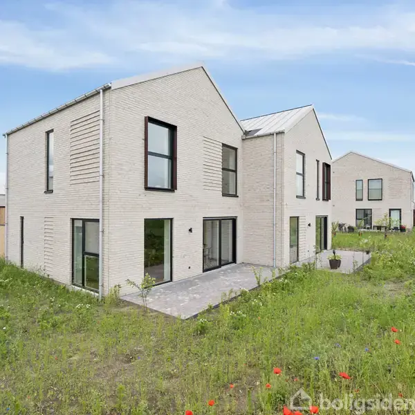 To moderne hvide murstenshuse står side om side på en græsmark med vilde blomster, under en blå himmel med få skyer.