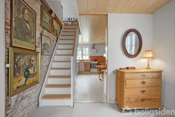 Trappe fører opad langs en mur med indrammede plakater. Til højre står en kommode med lampe og et ovalt spejl. I baggrunden ses en åbning til et køkken.