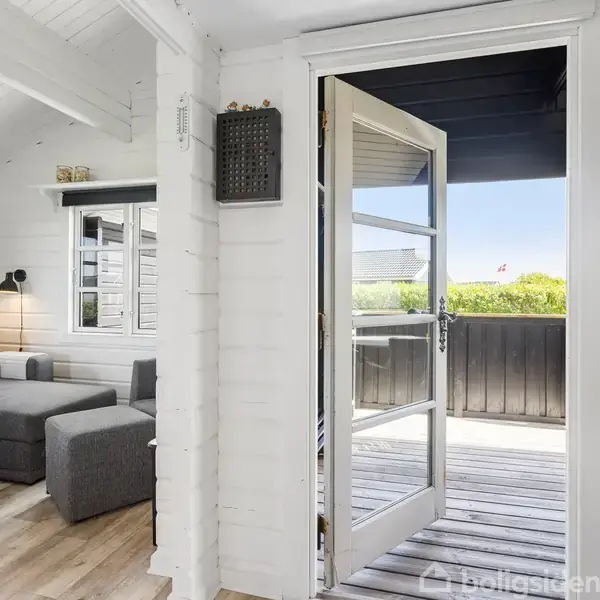 En åben dør fører fra en stue til en veranda. Stuen har en grå sofa, hvidt bord, og træmøbler, mens verandaen har udsigt til en grøn have og blå himmel.