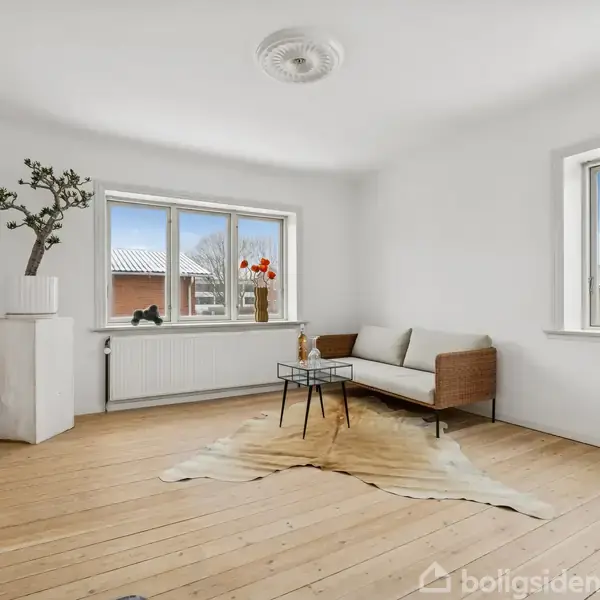 Sofamøbel står i et lyst rum med trægulv. Ved siden af er et lille bord og en vase med blomster. To vinduer lader lys trænge ind, og et tæppe ligger på gulvet.