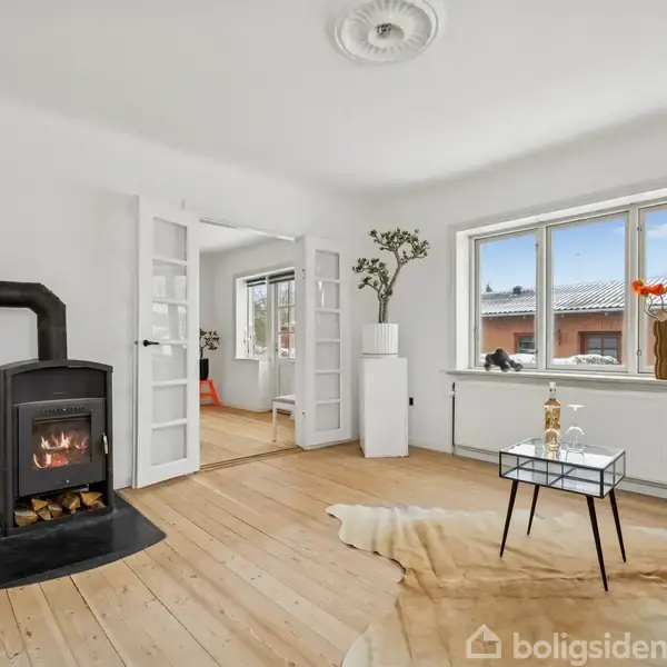 En brændeovn står i et hvidt stuehjørne, omgivet af trægulv og en sofa. Et bord med dekoration står ved et vindue med udsigt til et rødt hus.