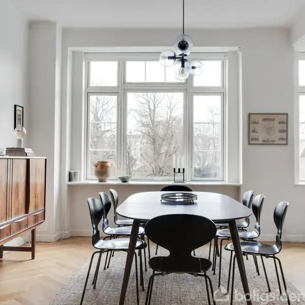Et spisebord er omgivet af otte sorte stole, placeret i et lyst rum med store vinduer. En træskænk med blomsterdekoration står op ad væggen, og en moderne lampe hænger fra loftet.