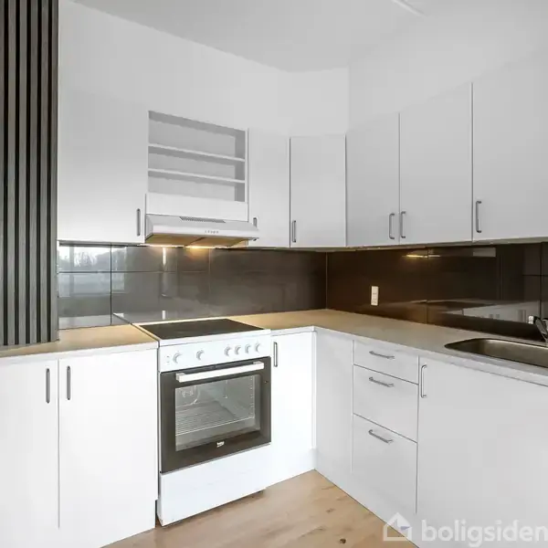 Et hvidt køkken med integreret komfur og ovn står i en hjørneopsætning. Køkkenskabe omkranser rummet, og der er en mørk bordplade med en vask.