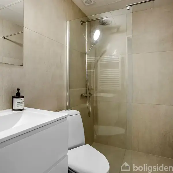 Håndvask med hvidt skab står ved siden af et toilet i et moderne badeværelse. Badeområdet har glasvæg og regnbruser. Vægge og gulv er flisebelagte i neutrale toner.