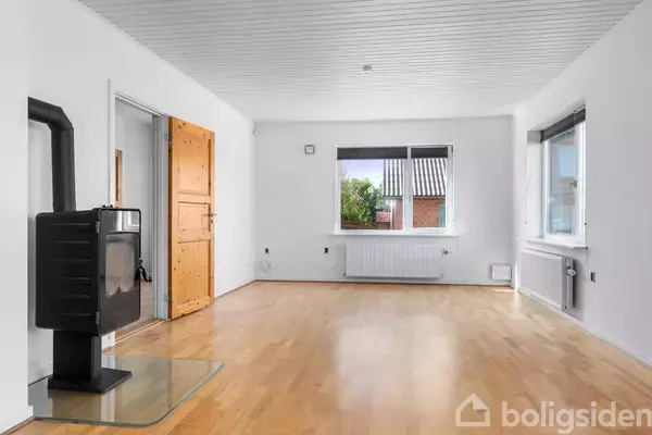 Stue med brændeovn står stille i et lyst rum med trægulv. To vinduer giver udsigt til en have. En trædør fører til et andet rum.