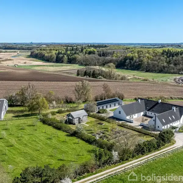 Hvidt bondehus med flere bygninger omgivet af grønne marker og skove. Et drivhus og en gårdsplads er nær huset, og der er en sø i baggrunden under en blå himmel.