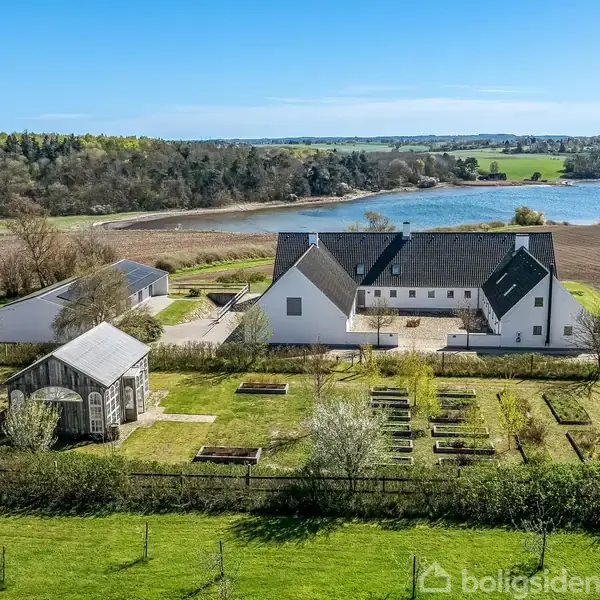 Landsted med hvid facade omgivet af marker og havebede. Lille drivhus ved siden af. Skov og sø i baggrunden. Klar blå himmel ovenover.