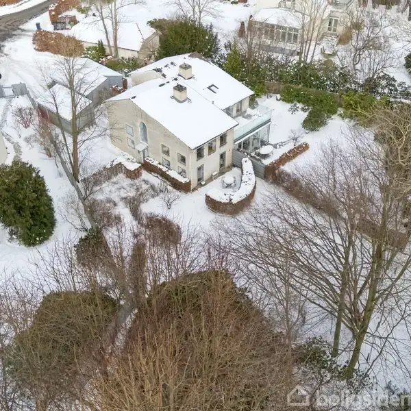 Et hus er dækket af sne, omgivet af træer og nærliggende bygninger. Det er en vinterlig scene i et boligområde, med et tydeligt lag sne på taget og jorden.