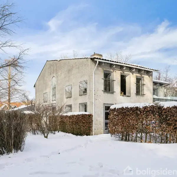 Et to-etagers hus står stille omgivet af sneklædte buske og træer, mens himlen er blå med få skyer.