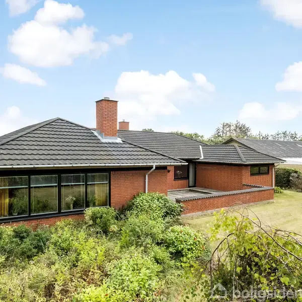 Et rødt murstenshus med sort tegltag står i en grøn have omkranset af buske og træer under en blå himmel med få hvide skyer.