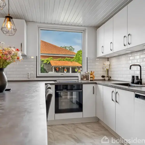 Et moderne hvide køkken har blomster i en vase på en marmorlignende bordplade. Rummet er oplyst af unikke pendellamper. Udsigten gennem vinduet viser et hus med rødt tag.