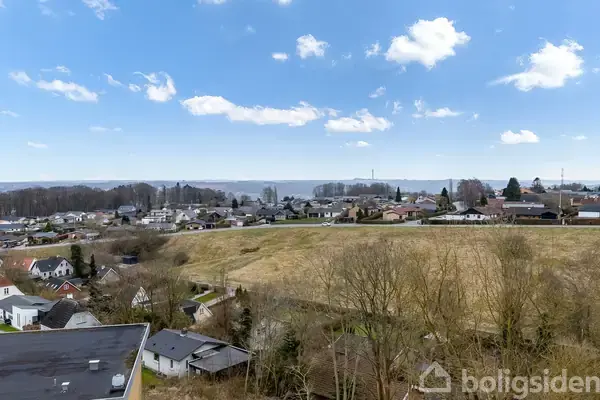 Udsigt over et boligområde med huse omkring en stor græsmark, mens himlen er skyet og blå. Landskabet strækker sig til en horisont med skov i baggrunden.