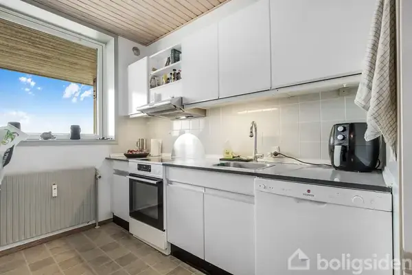 Et moderne køkken med hvide skabe, en komfur-ovn, og en opvaskemaskine. En kaffemaskine står på disken. Et stort vindue lader sollys trænge ind i rummet.