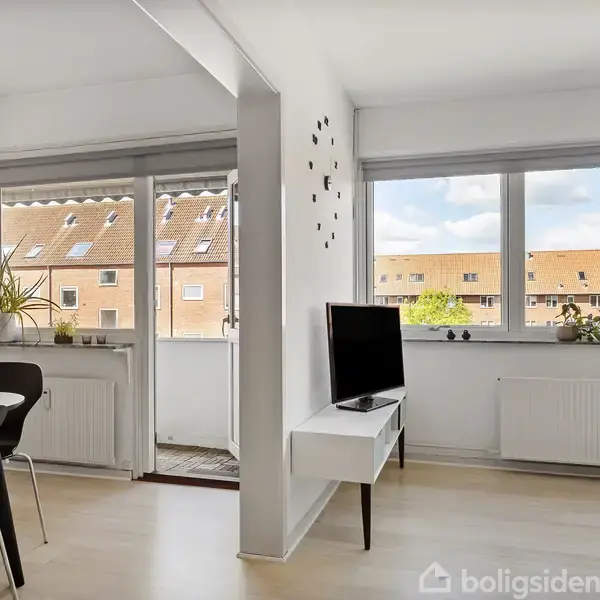 Stuen indeholder et fjernsyn på en hvid reol, vendt mod spisebordet med sorte stole. Sollys strømmer gennem store vinduer, udsigt til nabobygninger med røde tage.