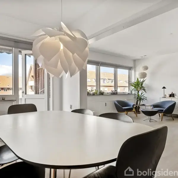 Ovalt spisebord med seks stole står i en lys stue. En unik lamel-lampe hænger over bordet. Store vinduer og planter pryder det moderne rum.