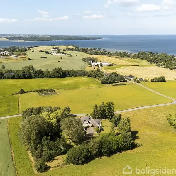 Luftfoto af landbrugsområde med marker og spredte gårde. Vejnet krydser landskabet. Baggrund viser kystlinje og hav. Træer og buske omkranser bygninger midt i billedets forgrund.