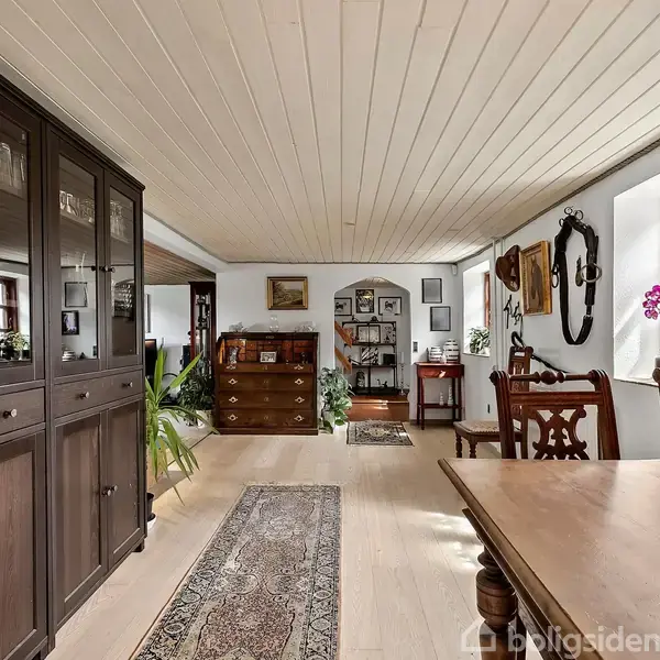 Et rummeligt spisestueområde med en mørk træskænk fyldt med glas. En træbord med dekorationer står omgivet af stole. Væggene har billeder og spejle; lyse træloft og gulv.