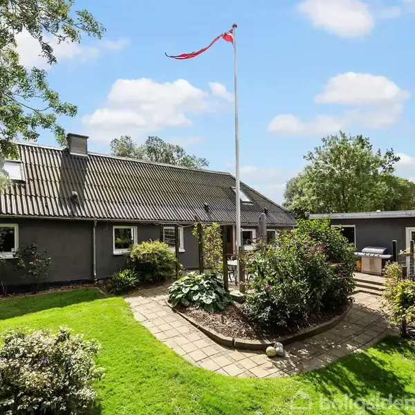 En gråmalet bygning med et skråtag står stille omgivet af en velholdt have. En dansk flagstang står i midten, omringet af frodige grønne planter og træer under en blå himmel.