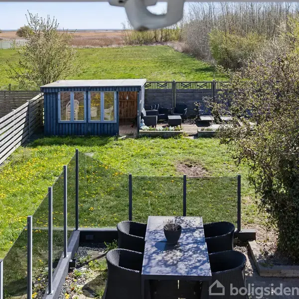 Terrassebord med sorte stole står på flisebelægning; i baggrunden ses en græsplæne med et blåt skur og havemøbler. Omgivet af hegn, enge og træer.