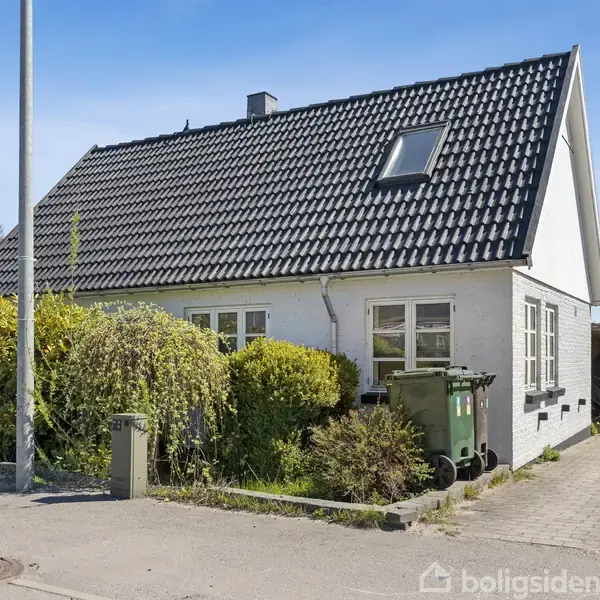 Et hvidt hus med sort tag står på en villavej. Foran huset er der en skraldespand, buske og en indkørsel. Himlen er klar og blå.