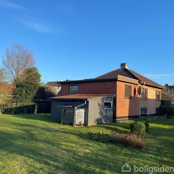 En rød murstensvilla med brunt tag står i en solrig have. Ved siden af huset er en grå skur. Græs og buske omgiver huset under en blå himmel.