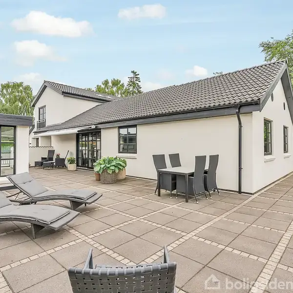 En hvidmalet villa med sort tag ligger i en solrig have. To liggestole og et bord med stole står på den flisebelagte terrasse. Grønne træer omkranser ejendommen.