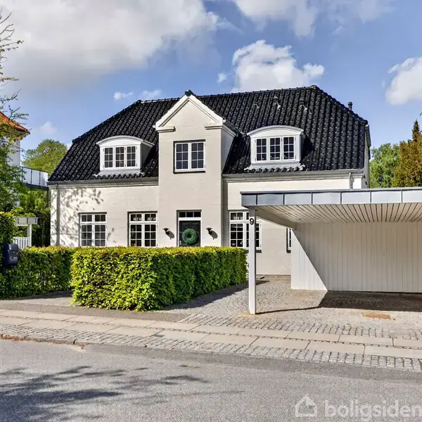Et hvidt hus med sort tegltag står bag en grøn hæk ved en rolig vej. En carport til højre giver skygge, omgivet af frodig vegetation.