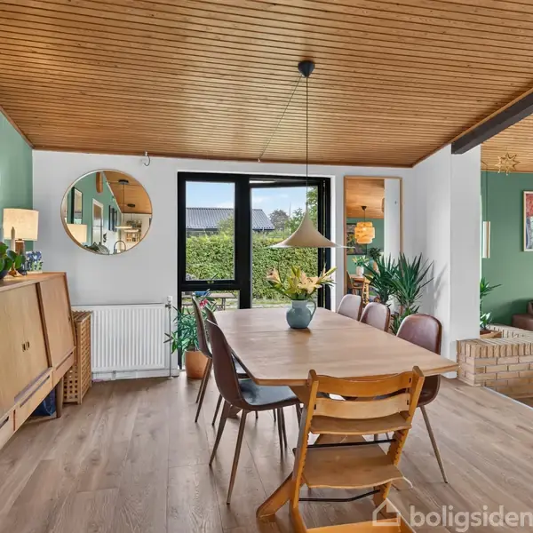 Et spisebord med stole står i et hyggeligt rum med træloft. En stor potteplante ved vinduet tilføjer grønt. Kunstværker pryder de grønne vægge. En sofa ses i baggrunden.