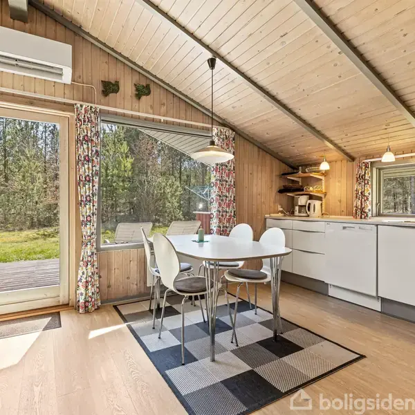 Rundt spisebord med stole står på et tæppe i et lyst køkken-alrum. Store vinduer viser udsigt til skov. Loftlampe hænger over bordet, og aircondition er placeret øverst.