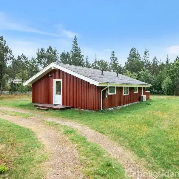 Rødt træhus står stille på en grøn græsplæne omgivet af høje træer. En grussti leder op til huset. Himlen er klar med lette skyer.