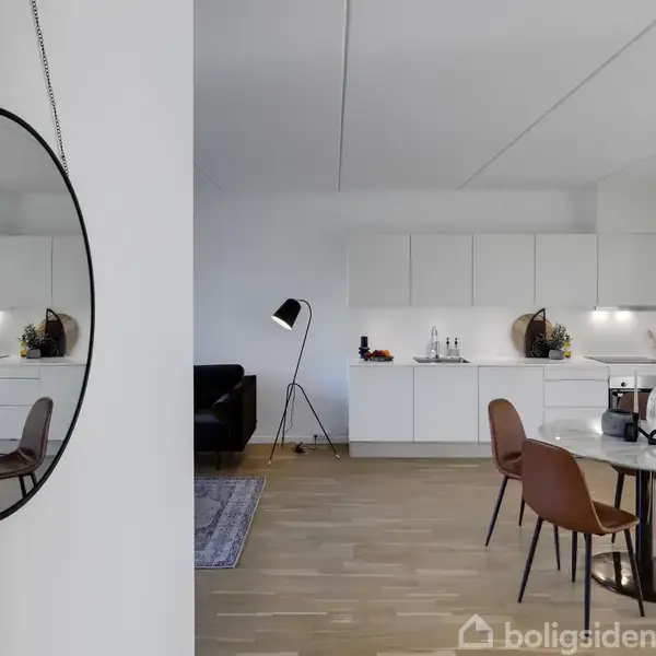 Et rundt spejl hænger på væggen, spejler spisebord og stole i et moderne køkken-alrum med hvide skabe og en sort gulvlampe.