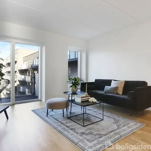 En stue med sort sofa og gulvlampe til højre. Et tæppe ligger under et bord med bøger og planter. Store vinduer viser en altan og nabobygninger udenfor.