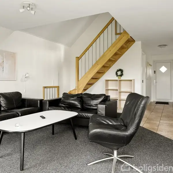 Sort læder sofaarrangement står i en stue med gråt gulvtæppe; en trætrappe fører opad i baggrunden, og vægge er dekoreret med kunstbilleder. Hvidt sofabord foran sofaen.