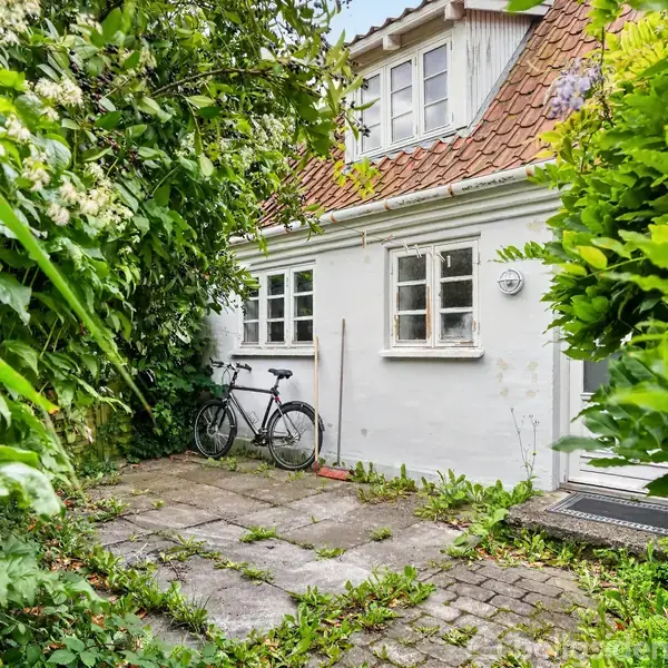 En cykel står op ad en hvid murstensvæg ved siden af en kost. Omgivet af tæt vegetation foran et hus med rødt tegltag og små vinduer.