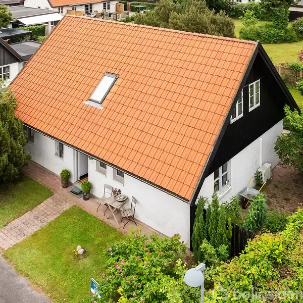 Et hvidt hus med rødt tegltag og en kvist, står ved en sti. En lille terrasse med bord og stole ses foran. Grønt buskads omgiver huset.